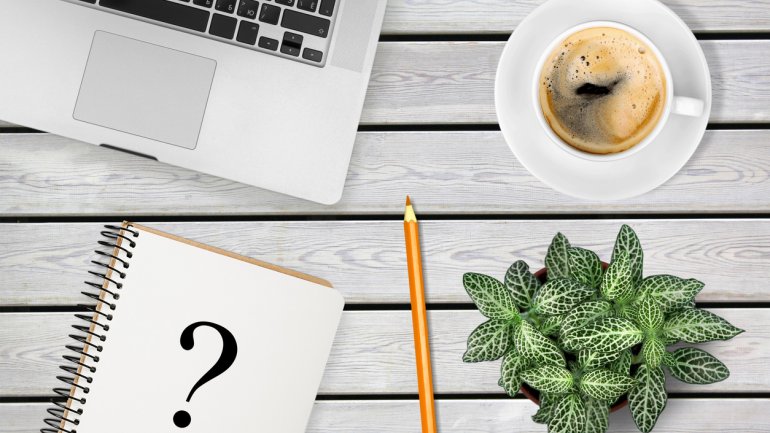 Laptop, Kaffee, eine Pflanze, ein Stift und ein Block mit einem Fragezeichen liegen auf dem Tisch