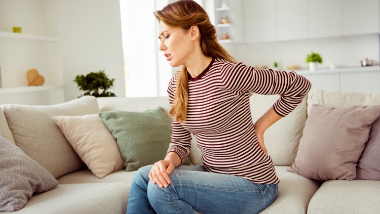 Frau sitzt auf dem Sofa und hat menstruationsbedingte Rückenschmerzen