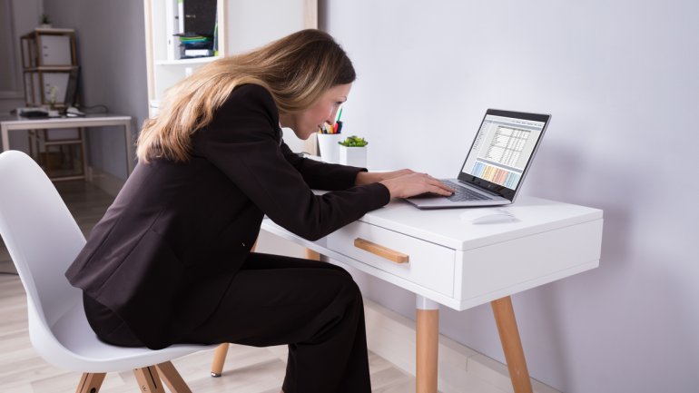 Frau sitzt schräge und verkrampft am Schreibtisch und beugt sich vor zum Laptop