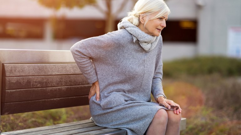 Eine ältere Frau sitzt auf einer Parkbank und hält ihre Hand an den Rücken
