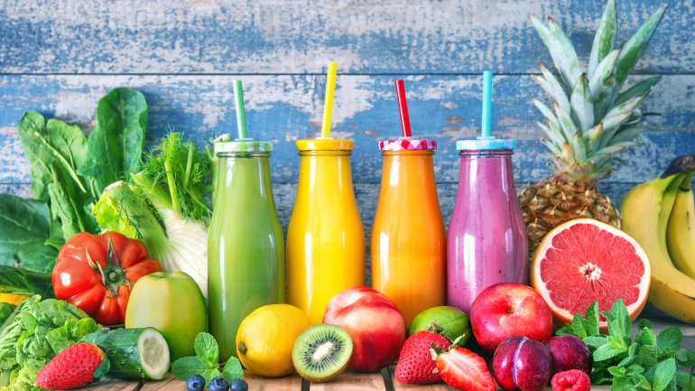 Vier Glasflaschen mit bunten Smoothies in Pink, Orange, Gelb und Grün zwischen Gemüse