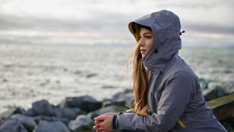 Eine junge Frau steht in einer Windjacke am Meer und hat die Kapuze auf