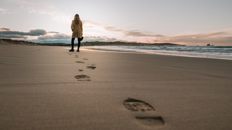 Ein Strand mit Schuhabdrücken, im Hintergrund steht eine Frau mit Blick aufs Meer